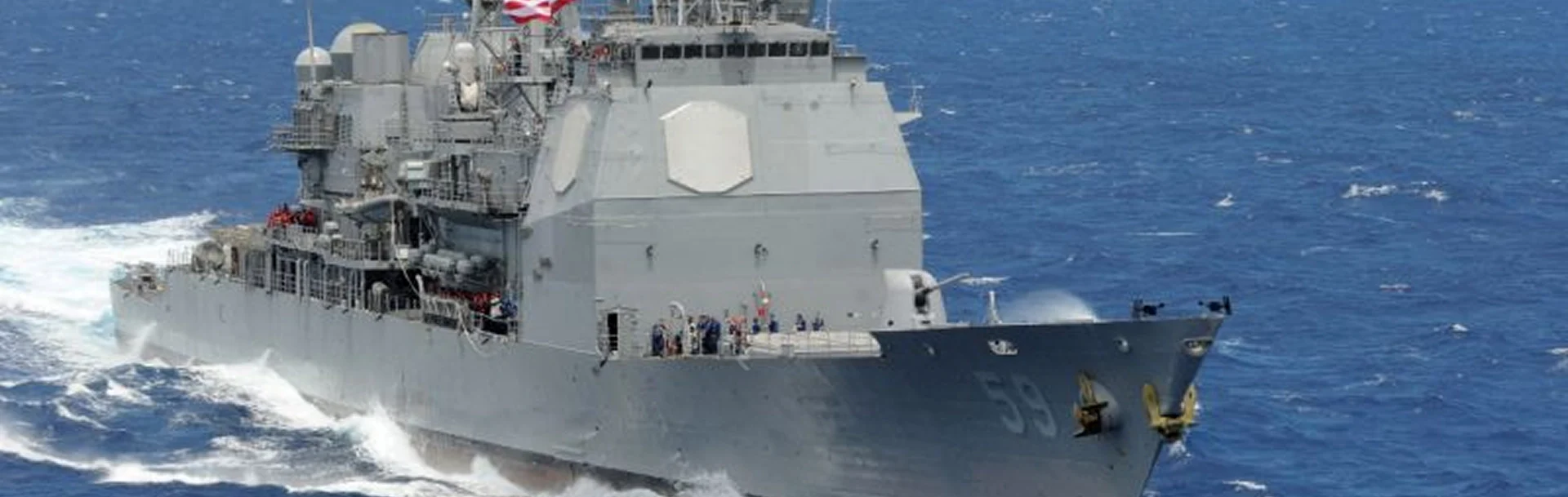 A naval ship navigates through ocean waves, showcasing its sleek gray hull and equipment against a backdrop of blue water.